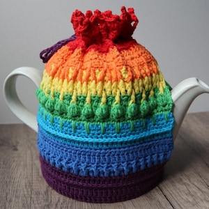 Op de afbeelding: Een kleurrijke, gehaakte theemuts in regenboogkleuren, waaronder rood, oranje, geel, groen, blauw en paars. De theemuts is ontworpen om over een witte theepot te passen, met een trekkoordsluiting aan de bovenkant.
