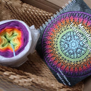 May include: A rainbow yarn skein and a crochet pattern book titled "Mandala 'Lasting Love'" by Wizard of Loops.