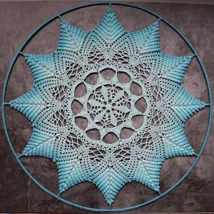 Op de afbeelding: Een grote, blauwe gehaakte doily met een stervormig ontwerp. De doily hangt aan een rond metalen frame.