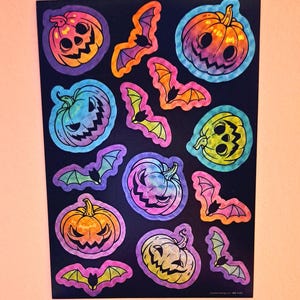 May include: A sheet of holographic stickers featuring a repeating pattern of rainbow colored pumpkins and bats. The stickers are on a black background.
