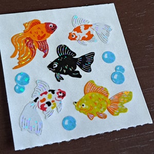 May include: A sheet of colorful goldfish stickers with iridescent glitter. The stickers feature four goldfish in various colors and patterns, including orange, red, black, and yellow. There are also four blue iridescent stickers that resemble bubbles.