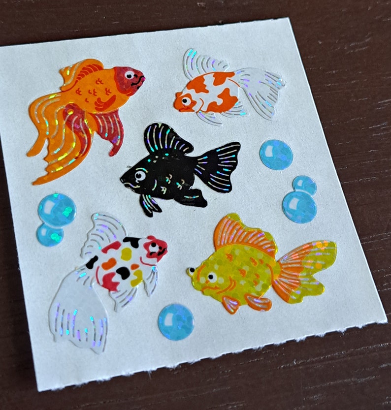 May include: A sheet of colorful goldfish stickers with iridescent glitter. The stickers feature four goldfish in various colors and patterns, including orange, red, black, and yellow. There are also four blue iridescent stickers that resemble bubbles.