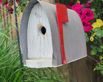 May include: A birdhouse designed to resemble a mailbox, featuring a weathered white wooden front with a circular entrance hole, a curved gray metal roof, and a bright red metal flag attached to the side, suspended from a black metal hook.