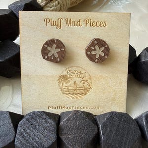 Peut inclure: Une paire de boucles d'oreilles à clous en bois brun foncé en forme de dollars des sables, fixées sur une carte de présentation carrée de couleur claire. La carte porte le texte Pluff Mud Pieces et un logo circulaire avec un palmier, un pont et un dauphin, ainsi que le site web PluffMudPieces.com.