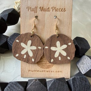Peut inclure: Une paire de boucles d'oreilles rondes en bois brun foncé avec un motif découpé de dollar des sables de couleur claire. Les boucles d'oreilles sont suspendues à des crochets en métal doré sur une carte en bois clair marquée Pluff Mud Pieces.