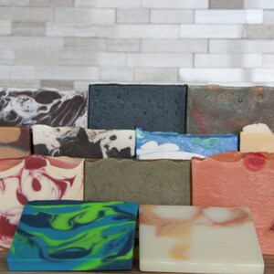 May include: A collection of handmade soaps in various colors and patterns. The soaps feature swirls, marbling, and layered designs in shades of blue, green, red, white, and brown. Some soaps have embedded decorative elements.