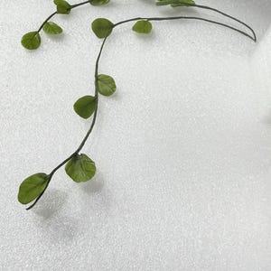 May include: Two artificial green vine stems with small, rounded leaves. The stems are dark green and curve gracefully, with leaves evenly spaced along their length. The leaves are a vibrant green color.