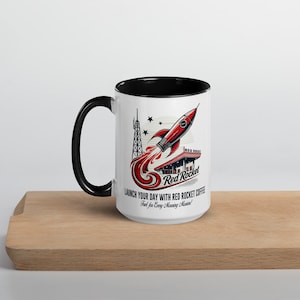 Może przedstawiać: Biały ceramiczny kubek do kawy z czarnym rantem i uchwytem. Kubek przedstawia retro ilustrację czerwonej rakiety startującej z czerwono-białego budynku z tekstem "Red Rocket" i "Launch your day with Red Rocket coffee. Fuel for every morning mission!"