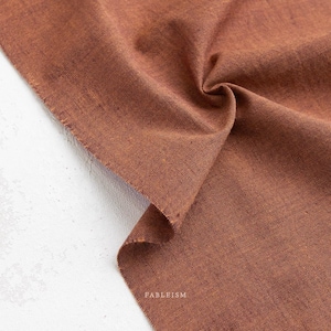 May include: Close-up of a folded piece of brown fabric with a textured surface. The fabric has a raw edge and the word "FABLEISM" is printed in white on the lower right corner. The fabric is on a white surface.