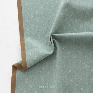Könnte beinhalten: Ein salbeigrüner Stoff mit einem sich wiederholenden Muster aus kleinen weißen gestickten Kreuzen. Das Textil hat eine markante braune Webkante an einer Seite und enthält das Wort FABLEISM, das unten mittig in Weiß auf den Stoff gedruckt ist.