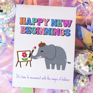 Op de afbeelding: Een wenskaart met de woorden "HAPPY NEW BEGINNINGS" in kleurrijke letters. Een illustratie van een grijze olifant met een baret die een bloem op een ezel schildert, is te zien. De tekst "Het is tijd om weer in contact te komen met de magie van hobby's" staat eronder.