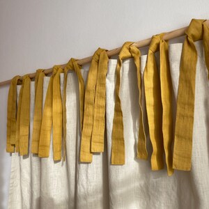 Puede incluir: Un par de cortinas de color blanco roto colgadas de una barra de madera clara. Las cortinas cuentan con lazos de tela de color amarillo mostaza anudados alrededor de la barra para sujetar los paneles, creando un aspecto fruncido y texturizado.