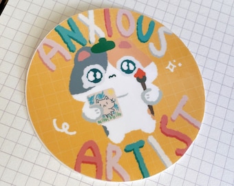 May include: A round sticker with a yellow background and a white cat holding a paintbrush and a picture. The text "ANXIOUS ARTIST" is written in colorful letters around the cat.