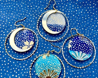 Può includere: Quattro paia di orecchini a cerchio con disegni celesti. Ogni orecchino presenta un cerchio color oro con un ciondolo circolare. I ciondoli mostrano motivi di luna e sole con sfondi a pois blu e bianchi.