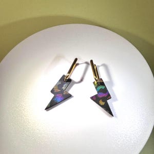 Op de afbeelding: Een paar zwarte bliksemvormige oorbellen met goudkleurige creolen. De bliksemschichten hebben een holografisch, iriserend patroon dat meerdere kleuren reflecteert. De oorbellen worden op een wit oppervlak gepresenteerd.