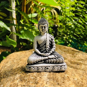 May include: A white ceramic statue of Buddha sitting in meditation pose. The statue is detailed with a patterned robe and a serene expression.