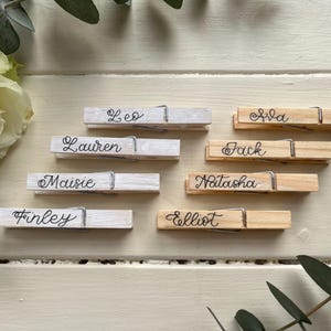Può includere: Mollette di legno con nomi scritti a mano: Leo, Lauren, Maisie, Finley, Ava, Jack, Natasha ed Elliot. Alcune sono verniciate di bianco, altre sono in legno naturale. Una rosa bianca e del verde sullo sfondo.