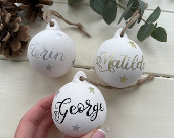 May include: Three white ceramic Christmas ornaments with gold and silver stars and personalized names: Erin, Matilda, and George.