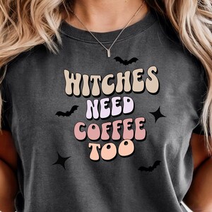 以下が含まれることがあります： レトロなフォントで「Witches Need Coffee Too」と書かれた濃い灰色のTシャツ。デザインにはコウモリと星が含まれています。このシャツはカジュアルで快適なスタイルです。