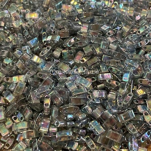 Pode incluir: Um grande monte de contas de sementes prateadas iridescentes. As contas são pequenas e retangulares com dois furos em cada extremidade.