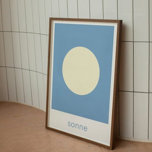 May include: A framed print with a light blue background and a large, off-white circle in the center. The word "sonne" is printed in blue at the bottom of the print.