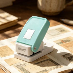 Puede incluir: Perforadora de papel con forma de sello, verde menta y blanca, con el nombre de la marca "KUHINO" impreso en el lateral. La perforadora está sobre una página de sellos antiguos.