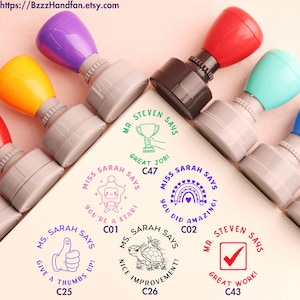 Pode incluir: Uma coleção de carimbos auto-entintados coloridos com vários desenhos e frases. Os carimbos incluem frases como "Bom trabalho!", "Você é uma estrela!" e "Joinha!" Cada carimbo tem uma alça de cor diferente.