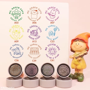 Könnte beinhalten: Eine Sammlung farbenfroher Selbstfärbestempel mit verschiedenen Designs und Sätzen wie „Great Job!“ und „Excellent Work!“. Die Stempel zeigen Cartoon-Charaktere und ermutigende Botschaften. Im Hintergrund befindet sich eine dekorative Zwergfigur.