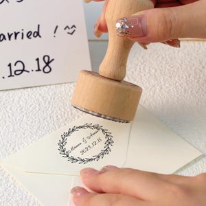 Könnte beinhalten: Ein Holzstempel mit kreisförmigem Design wird auf einen weißen Umschlag gedrückt. Der Stempel zeigt ein Kranzdesign mit den Namen "Manna & Annie" und dem Datum "2023.12.18". Der Stempel wird verwendet, um den Umschlag zu personalisieren.