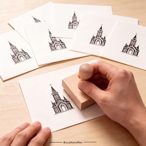 May include: A wooden rubber stamp with a handle, used to imprint a black ink illustration of a church building onto white paper cards. Several identical cards featuring the same black church design are arranged in the background.