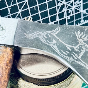 May include: A metal hatchet with a wooden handle featuring an engraved deer on the blade and the text Ironside Knife & Axe with a circular logo. The axe head is silver and dark grey, showing a detailed etched illustration of a leaping buck.