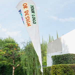 May include: White feather flag with the text "YOUR LOGO OR DESIGN" in colorful letters. The flag is displayed outdoors against a backdrop of green trees and a white building.
