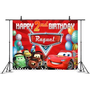 May include: A red backdrop with the text "HAPPY 2nd BIRTHDAY" and the name "Raquel". The backdrop features cartoon cars and balloons. The backdrop is supported by a black stand.