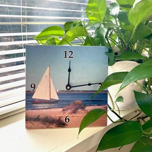 May include: Square wall clock featuring a coastal scene with a sailboat on the water. The clock face has black hands and large black numerals at 12, 6, and 9. The clock is displayed on a white surface with green plants in the background.