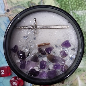 May include: A black candle container filled with white wax, amethyst crystals, and silver glitter. A silver sword charm rests on top. Red dice are scattered around the candle, on a map.