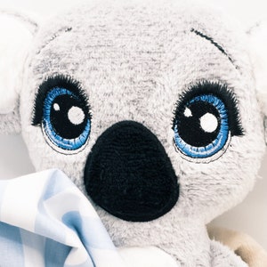 以下が含まれることがあります： 青い目と黒い鼻をした灰色と白のぬいぐるみコアラ。コアラは青と白のチェック柄の毛布を持っています。画像の右下隅に「KrossMB Designs」というテキストが表示されています。画像は「13 cm x 18 cm HOOP」とラベル付けされています。