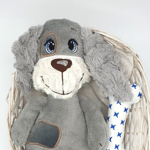 May include: A grey plush dog toy with blue eyes and a brown nose, sitting in a white wicker basket with a white and blue patterned bone toy. The text "KrosnUB Designs" is visible at the bottom of the image.
