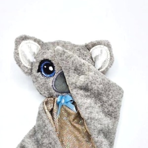 May include: A grey plush koala with a blue eye and a gold sparkly blanket. The koala is wearing a blue bow. The blanket is wrapped around the koala like a cape. The text "KrossMB Designs" is written in black at the bottom of the image.