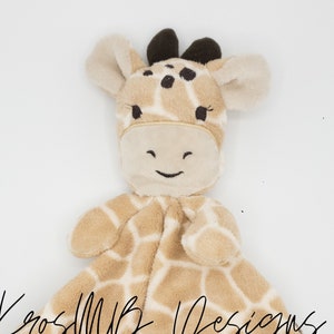 Puede incluir: Una manta de seguridad de peluche de jirafa de color marrón claro y marrón con una sonrisa bordada en marrón. La manta tiene un borde marrón y un patrón de jirafa.