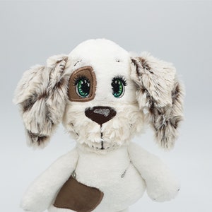 Puede incluir: Juguete de peluche blanco en forma de perro con detalles de piel marrón, ojos verdes y nariz marrón. El perro está de pie sobre una base negra con el texto "KrosnUB Designs" escrito en blanco.