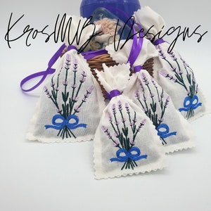 Puede incluir: Cinco pequeñas bolsas de tela blanca con bordados de lavanda y detalles de cinta azul. Las bolsas están dispuestas alrededor de un recipiente azul con una cesta de mimbre debajo. El texto "KrosMB Designs" es visible en la esquina superior derecha de la imagen.