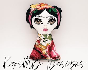 May include: A handmade doll with black hair, a floral crown, and a colorful embroidered dress. The doll has brown eyes and pink lips. The doll is sitting on a white background.