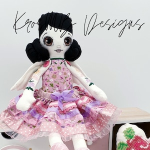 May include: A handmade doll with black hair, large eyes, and a pink and white dress. The doll is sitting on a white shelf with a rainbow and stars design. The doll is wearing pink shoes.