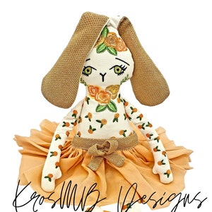 May include: A handmade embroidered bunny doll wearing a peach-colored tulle skirt and brown fabric shoes. The bunny has a white face with embroidered green eyes, a brown nose, and a floral pattern on its body.