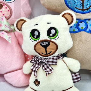 May include: A white plush teddy bear with brown embroidered ears, a brown nose, and large green eyes. The bear is wearing a brown and white checkered bow tie. The bear is sitting on a white surface.