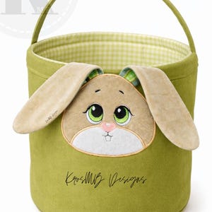 May include: A green Easter basket with a bunny face applique. The basket has a handle and a gingham lining. The bunny face features large green eyes, a pink nose, and tan ears. The text "KrosMB Designs" is on the basket.