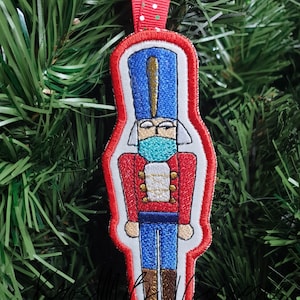 Op de afbeelding: Een rode en witte kerstversiering van vilt met een notenkraker die een blauwe hoed en een blauw en groen mondkapje draagt. De versiering heeft het jaar 2020 geborduurd op de onderkant.