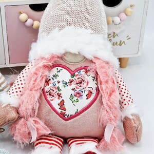May include: A pink and white gnome doll with a heart-shaped floral patch on its chest. The gnome is wearing a pink and white striped hat with a white pom-pom on top. The gnome is sitting on a wooden surface with a pink and white garland behind it.