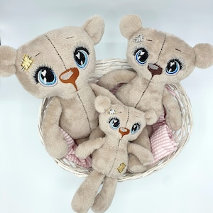 May include: Three plush teddy bears with embroidered eyes and patches, sitting in a white wicker basket. The bears are beige and have a distressed look.