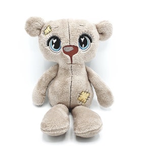 May include: A beige plush teddy bear with large blue eyes and embroidered details. The bear has patches sewn on its body and a stitched nose.